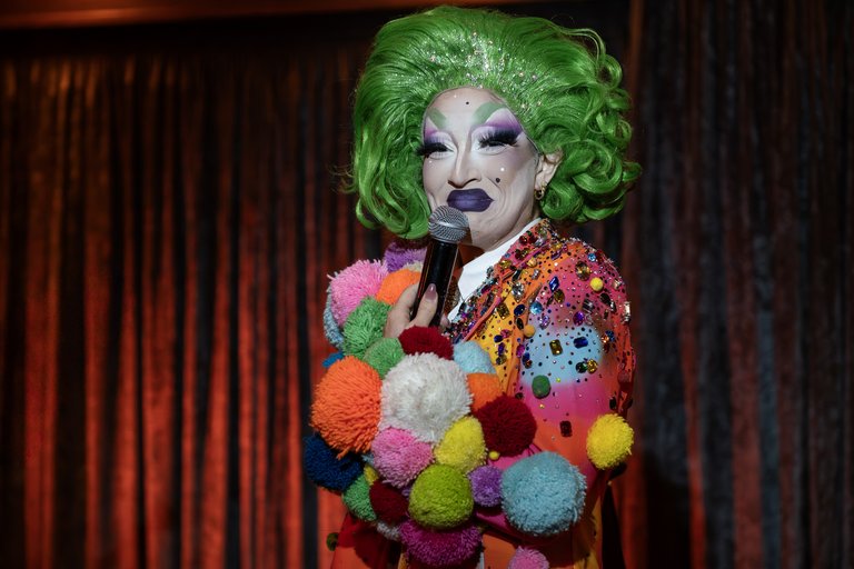 .	Sideshow nut performance at Liberty Festival 2025. Photo by Samuel Dore. A drag queen performer, Sideshow nut is shown on a stage, with red velvet curtains closed behind them. They are shown from t