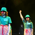 Two young people stand on stage wearing a green hat, green t-shirt, a red & white checkered apron and a red clown nose. Their right arm is lifted into the air and they are clicking.