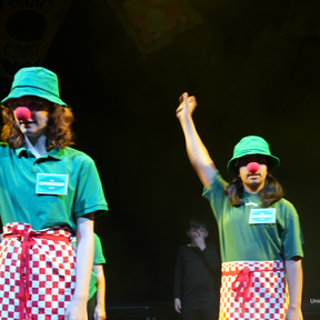Two young people stand on stage wearing a green hat, green t-shirt, a red & white checkered apron and a red clown nose. Their right arm is lifted into the air and they are clicking.