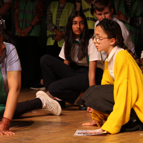 Two young people sit on the floor looking at each other with slight concern.